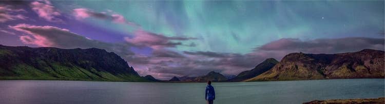 A person stands at the edge of a mountain lake beneath an aurora-lit night sky
